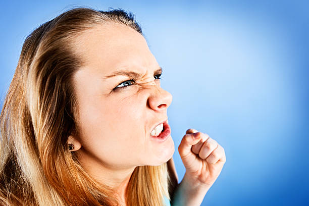 Furious blonde teenager shakes her fists and grimaces angrily An angry blonde teenage girl grimaces and shakes her fist at someone or something unseen. Blue background with copy space. punching one person shaking fist fist stock pictures, royalty-free photos & images