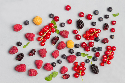 Still life of multi-colored berries on gray background. Red currants, raspberries red and yellow, blueberries, green mint. Flat lay