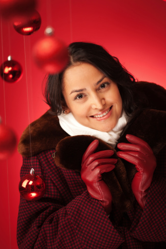 Subject: Vertical view of a beautiful, long-haired, Hispanic woman wearing a warm winter outfit of an old-fashioned, checked overcoat with fur collar, a white neck scarf, and red leather gloves. She holds up her collar for warmth and protection, and smiles for the camera in a Christmas theme red background setting. Red ball Christmas ornaments hang in the foreground.