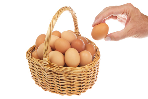 Hand places single egg to stack of them in cute wicker basket with a handle on white background