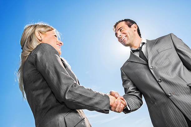 Cheerful blonde businesswoman shaking hands with her mid adult male colleague against the sky.