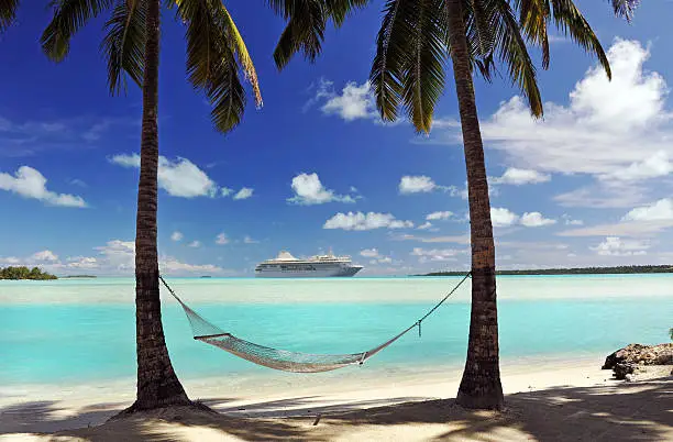 A cruise  ship passes an idyllic tropical island in the South Pacific with hammock and palm trees in semi-silhouette