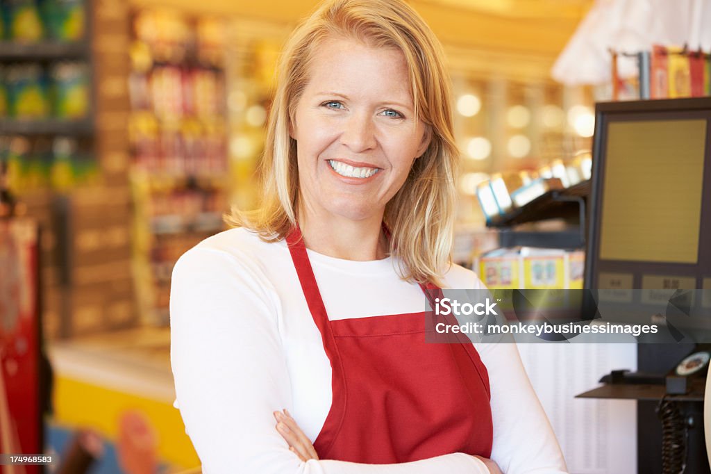 An older female cashier at a grocery store checkout Female Cashier At Supermarket Checkout Smiling To Camera Cashier Stock Photo