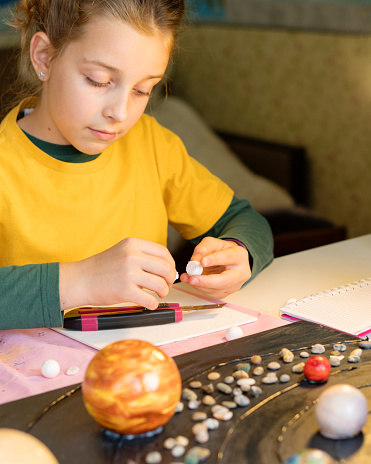 A diligent girl in a yellow shirt meticulously measuring and cutting, crafting a space-themed project with various beads and a notebook nearby