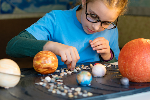 A young girl with glasses, engrossed in setting up a solar system display, carefully places planets and pebbles on a table.