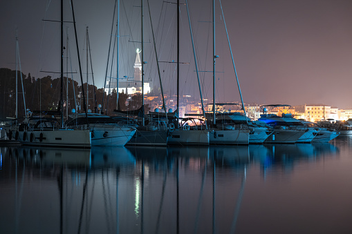 A view of the Saint Eufemia church and bell tower in the old city core and boats moored in the Adriatic sea next to buildings at night in Rovinj