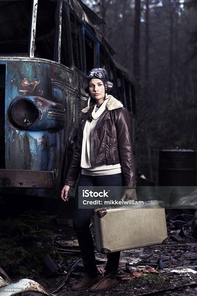 Female aviator travels with suitcase through junkyard Young woman in retro aviator outfit walks through a junkyard. She is holding an old suitcase. Bomber Jacket Stock Photo