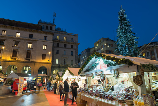 Varese, Italy - December 14, 2018: typical christmas market with lights and big Christmas tree in a city center at dusk. Varese city, square Monte Grappa, northern Italy. Varese is also called city garden for the presence of many parks and gardens. The town is famous for the Sacro Monte of Varese in 2003 entered from UNESCO in list of World Heritage. For Varese Lake famous because it hosts rowing competitions of national, European and world level. In the lake there is the Islet Virginia, an important Prehistoric pile dwellings around the Alps, from 2011, UNESCO Site