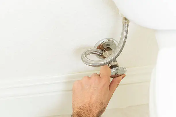 A hand turning a water shutoff valve mounted to a wall next to a toilet.
