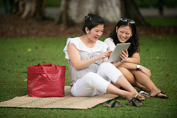 Women at the park A pregnant Asian woman with her sister reading and sharing a tablet at a park. beach mat stock pictures, royalty-free photos & images