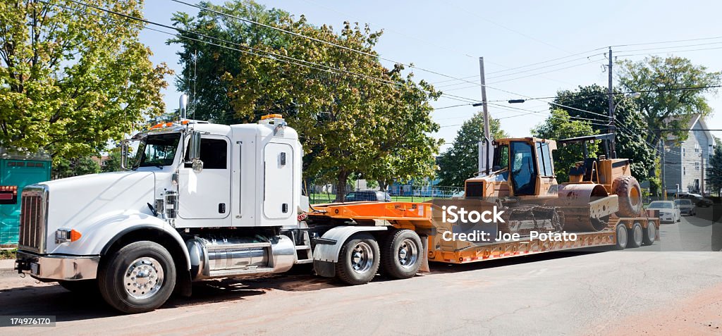Machinery Transport Semi, flatbed, and heavy machinery on residential street. Horizontal. Backhoe Stock Photo