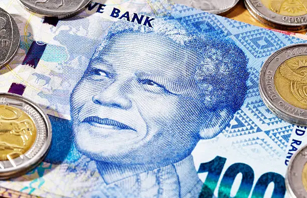 Close-up of a new South African One Hundred Rand banknote, featuring the smiling face of iconic statesman Nelson Mandela, with Five Rand and Two Rand coins scattered nearby. 