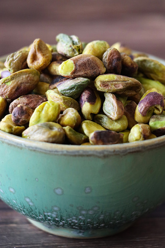 Stock photo showing close-up view of shelled pistachio nuts piled high in a green dish against a woodgrain background. Raw pistachios are considered to be a very healthy snack food and are high in vitamin B6, potassium, antioxidants and protein, boasting a list of health benefits and may aid weight loss.