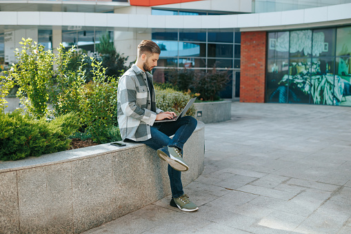 This portrait of a bearded young man working on a laptop outdoors exudes positivity and authenticity. With his stylish appearance and a shirt that adds to his fashionable look,