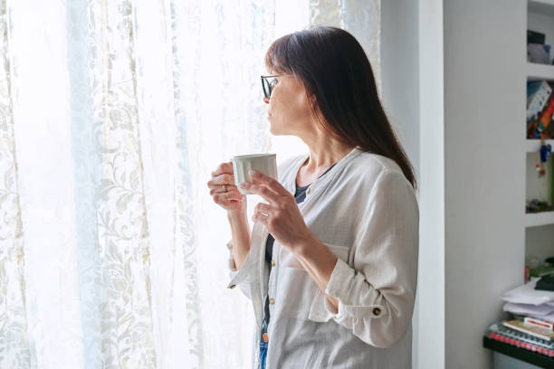 istock Serious middle-aged woman looking out window, with cup in hands 1749845243