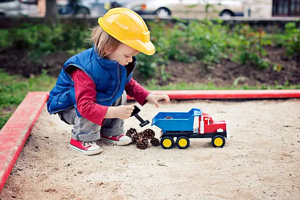 "Two years old boy, wearing yellow construction helmet and blue waistcoat, crouching in the sandbox and playing with toy truck and with pine cones. Very shallow dof, focus on the boy's right hand.Similar:"