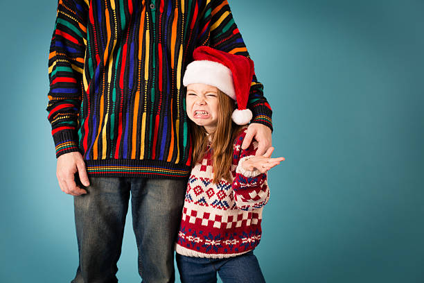Little Girl Making Face at Dad, Both Wearing Ugly Sweaters "Color image of a little girl with Santa hat, making a face at the ugly sweaters she and her dad are wearing." ugliness photos stock pictures, royalty-free photos & images