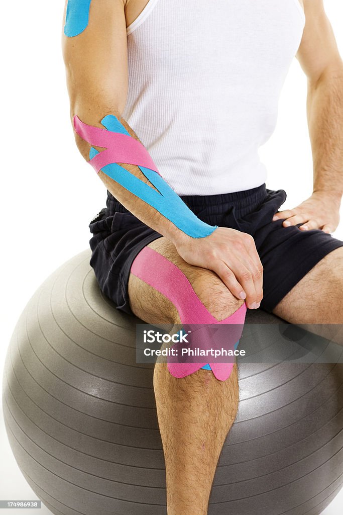 physical therapy Close up of a young man wearing sports clothing. Athlete with therapeutic tape around his arm and leg. Kinesiotape is used for prevention and treatment in competitive sports. Aching knee. Studio shot. High key. Isolated on white background. Elastic Therapeutic Tape Stock Photo