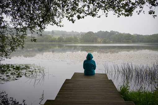 Rear view of silhouette of woman with blue raincoat sitting on the wooden pier in front of the lake on a rainy day. Concept of rural life, nature and landscape.