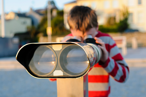 Boy in striped top looks through binoculars/telescope at a seafront promenade