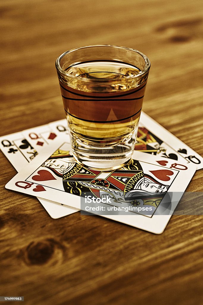 Whisky Queens Shot glass of whiskey resting atop four queen playing cards, standard classic design, queen of hearts prominently angled, textured wood surface. Shallow depth of field with selective focus on foreground card details. Fine grain added. Whiskey Stock Photo