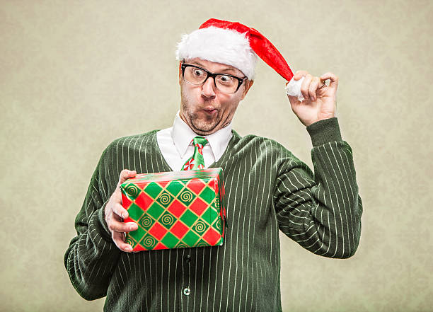 Funny Gift Exchange for the Holidays Man in Santa Hat A dorky dad in cardigan sweater holding holiday christmas gift. He is wearing a santa hat and a holiday festive neck tie.  He has a funny excited look on this face. christmas nerd sweater cardigan stock pictures, royalty-free photos & images