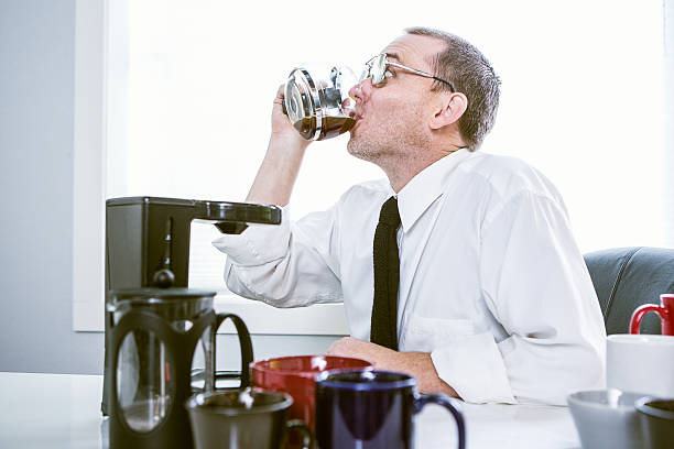 Coffee Troubles A man sits in his office cubicle surrounded by coffee makers and mugs, a hyper expression on his face from all the caffeine.  He drinks straight out of the coffee brewer carafe.  Horizontal. coffee addict stock pictures, royalty-free photos & images