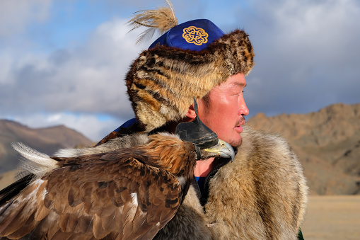 Side profile of a Kazakh eagle hunter with his hooded eagle at the Sagsai Golden Eagle Festival held on 17th-18th September 2023, near the small town of Bayan-Ulgii province district of Western Mongolia. Set in the Altai Mountain range, the nomadic eagle hunters, dressed in traditional fur and embroidered clothing, gather to compete with their eagles, in various competitions that show the skill and bond between the hunter and their eagle.