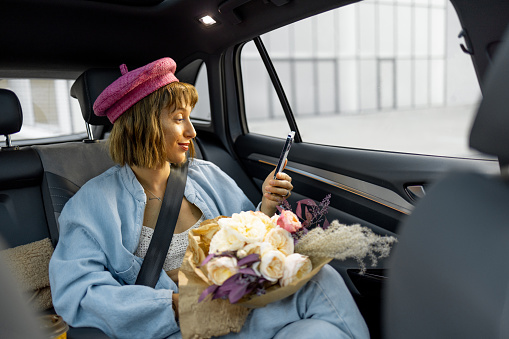 Young stylish woman uses smart phone while sitting with a flower bouquet on backseat of car on the go. Concept of transportation and urban lifestyle