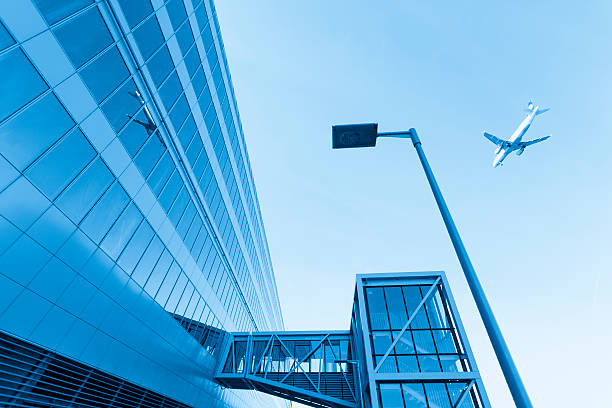 Airplane and Frankfurt airport building exterior Frankfurt airport scene with airplane flying over head. Lamp post next to building. Newer architecture. Airport blue hue. Upward sky angle. The sky is blue as well. Two buildings, lamp post, and airplane. Foreign airport. Sky view with mirrored building. Flying plane above.  frankfurt international airport stock pictures, royalty-free photos & images