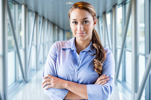 Young Hispanic Businesswoman in Bright Corridor Young Hispanic businesswoman in blue, standing with arms folded in a long, bright corridor of glass and steel. general manager stock pictures, royalty-free photos & images