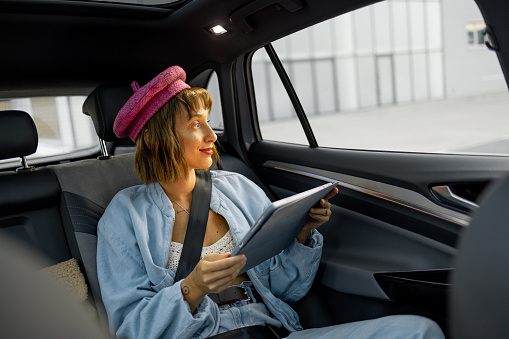 Young stylish woman uses touchpad, doing some creative work while sitting on backseat at car on the go. Concept of transportation and modern digital lifestyle