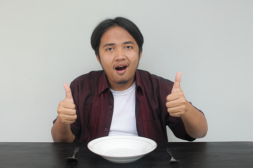 Asian young man in red shirt sitting looking in front of empty plate and showing wow and happy expression and thumbs up gesture. Eat concept.