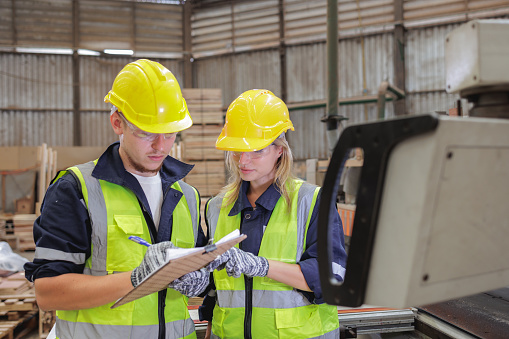 A young Caucasian male and female inspect a checklist at a warehouse, ensuring protocol compliance. Their diligent oversight underscores the importance of safety and precision in industry