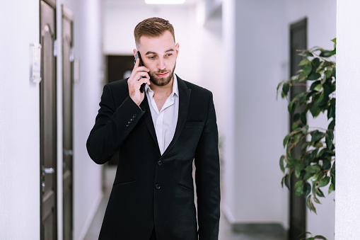 Experienced proud boss in black suit talking on telephone device in office corridor
