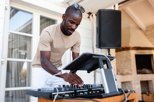A black adult male DJ playing and mixing music during an outdoor summer party at a beach house. He is wearing casual summer clothes and looks focused.