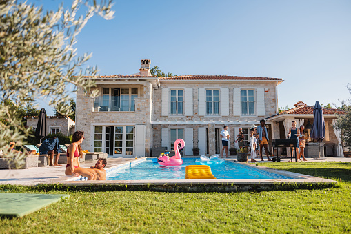 Diverse group of adult friends socializing by the pool during a fun pool party. They are enjoying hanging out and relaxing by the pool. They are wearing their colourful bikinis. The group of friends is located in the backyard of a beautiful Mediterranean style villa. Making new memories with friends during summer vacations in Croatia.