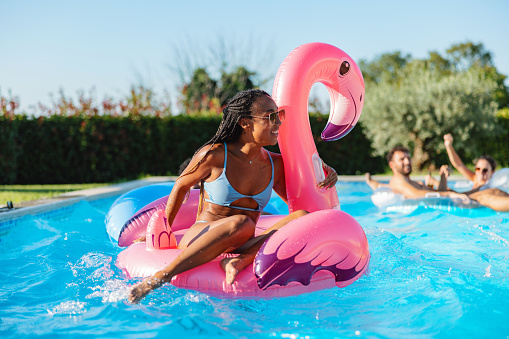 Happy black young woman lying on a flamingo float during a pool party. She is smiling and enjoying her time playing in the pool with friends. She is wearing a baby blue bikini and some sunglasses. Fun summer weekend with friends.