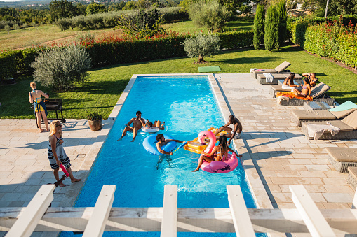 Multiracial group of diverse friends hanging out by the pool of a fancy villa. They are having a pool party in the summertime. The friends swimming in the pool and lying on the pool floats. The weather is sunny and perfect for sunbathing.