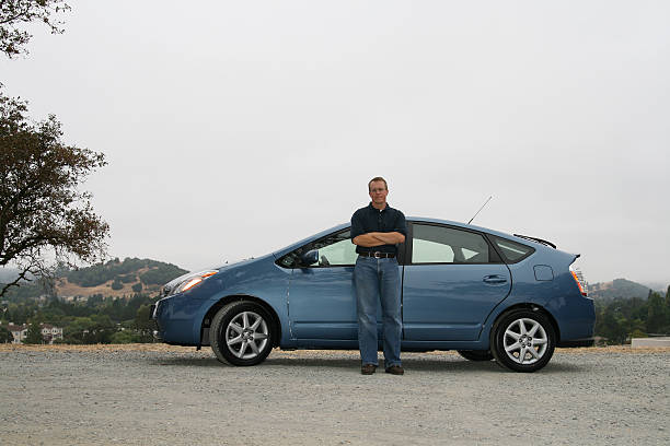 A man with glasses in jeans infront of a brand new hybrid (gas/electric) car. The car is blue and parked on gravel. Taken on an overcast day. It's a \