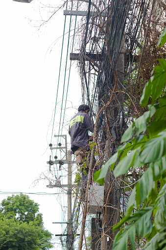 Thai telecommunication electrician on ladder is working on street overhead cables in a street in residential district of Bangkok Ladorao. Man is working for company AIS. He has logo and brand name on his jacket.