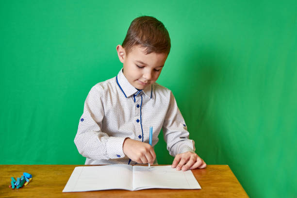 Cute boy draws with a pen in a notebook sitting at the table on a green background.