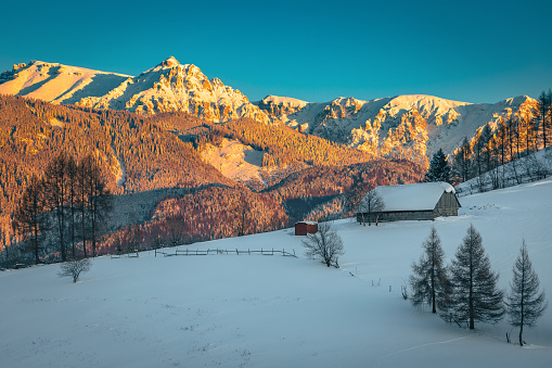 Beautiful winter landscape with snow covered wooden hut on the slope and majestic high mountains in background at sunset, Bucegi mountains, Carpathians, Romania, Europe