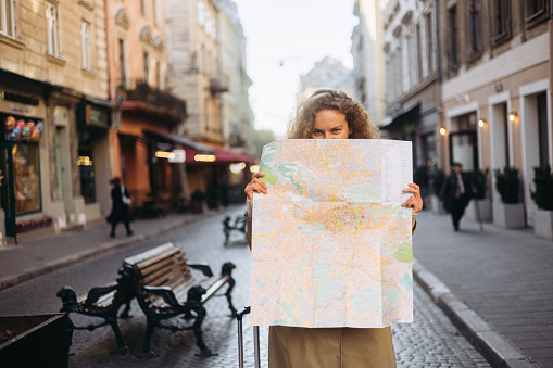 The radiant smile of a curly-haired woman as she navigates her way through an ancient European city during her autumn vacation, holding a tourist map