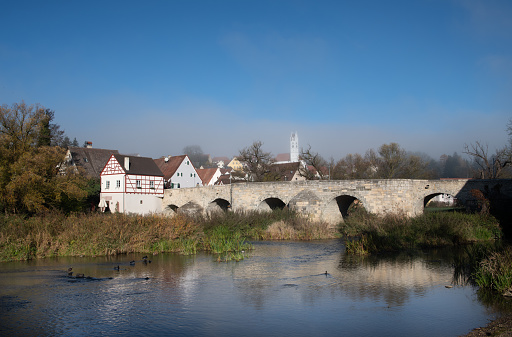 In the city of Harburg, an old stone bridge leads over the Wörnitz. The bridge is reflected in the water. Mist rises from the water.