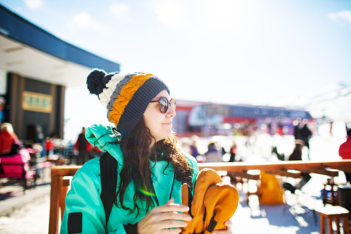 Young adult woman having fun at ski resort at sunny day, wearing knit hat, sunglasses and ski gloves