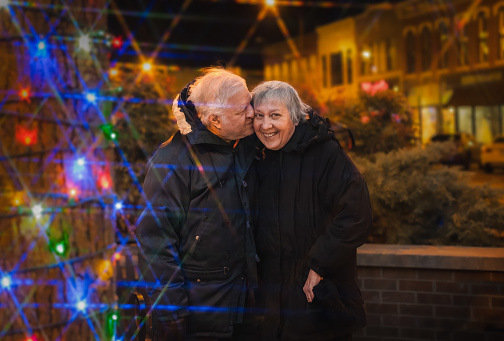 Senior couple embraces outdoors on street decorated for Christmas; charismas lights all around them