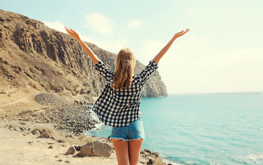 Back view, happy cheerful young woman raising her hands up on sea coast with mountain background, Tenerife, Canary Islands, Spain