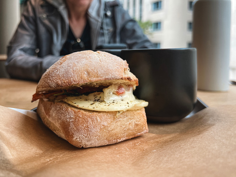 Delicious egg breakfast sandwich on a Ciabatta bun. Breakfast or brunch with a cup of coffee in a black coffee mug. A friend sits across the table looking at her mobile phone. Two friends sit across a wooden table from each other.