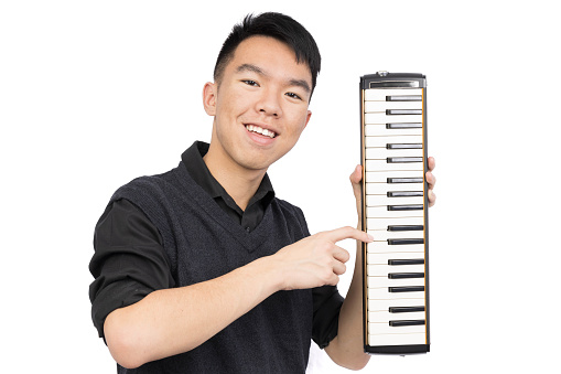 A young male is smiling and looking at the camera, holding up a melodica instrument and pressing a key on it. He is wearing a black shirt and a vest sweater. He is in a studio with a isolated white background.